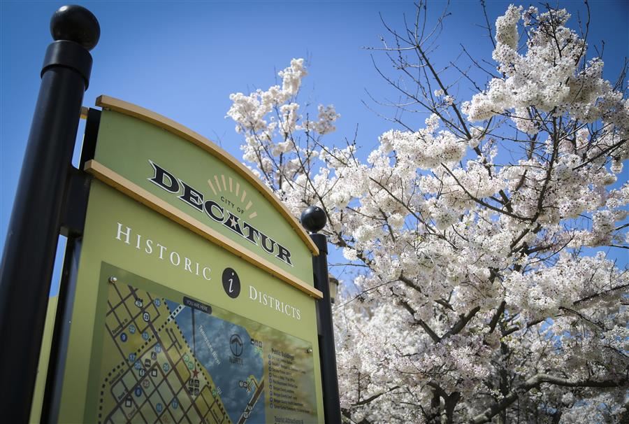 An informational sign reading "City of Decatur Historic Districts." Behind the sign is a blue sky and a dogwood tree in bloom